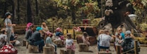 A group of children and adults are seated on tree stumps in an outdoor forest setting, listening attentively to a storyteller or performer. They are gathered around an area with a large, whimsical tree sculpture with a face, which appears to be part of a magical or fantasy-themed environment. The scene is surrounded by lush greenery and playful decorations like giant mushrooms.