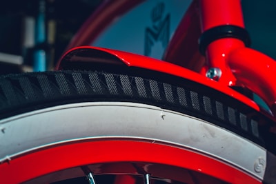 A close-up view of a bright red bicycle tire and frame section. The tire features a black tread pattern and is accented by a white rim. The red frame includes a shiny metallic bolt, adding detail to the design.