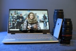 A silver laptop is displayed with its screen on, showing a wallpaper featuring a person in a checkered shirt holding a camera. The person is surrounded by cyclists in a blurred background. On either side of the laptop, camera lenses and a silver smartphone are placed, giving a tech and photography theme. The surface is covered with a textured blue material, providing a soft contrast.