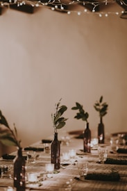 A long dining table set with minimalistic decor, featuring dark glass bottles used as vases containing small leafy branches. The table is adorned with small, lit candles adding a warm ambiance. Fairy lights are draped above, providing a cozy and intimate environment.