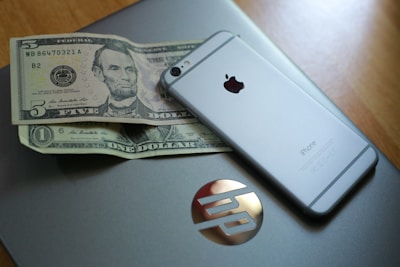 An HP laptop is placed on a wooden surface with two US dollar bills and an iPhone on top of it. The iPhone is positioned diagonally across the laptop.