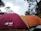 A campsite scene features two tents, one maroon and another orange, set up under a partly cloudy sky. Surrounding the tents are tall trees, adding a natural setting to the site. A pair of shoes is partially visible on the ground, suggesting recent human activity.