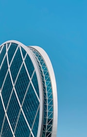 An architectural structure with a large, circular building featuring a grid of crisscrossing diagonal lines made of glass and metal. The building has a modern design and stands against a clear blue sky, highlighting its futuristic aesthetics.