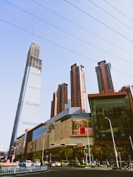 A cityscape featuring a collection of modern skyscrapers, with one prominent building under construction. A large commercial building is in the foreground, displaying an electronic billboard with an advertisement. Numerous vehicles and pedestrians are visible on the street, indicating urban activity.