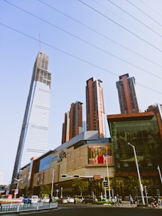 A cityscape featuring a collection of modern skyscrapers, with one prominent building under construction. A large commercial building is in the foreground, displaying an electronic billboard with an advertisement. Numerous vehicles and pedestrians are visible on the street, indicating urban activity.