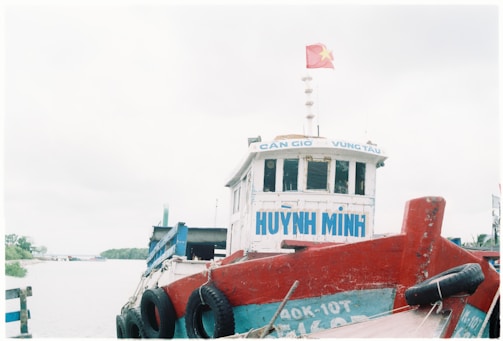 A large boat with a red hull and blue accents is docked near a waterway. The name 'HUYNH MINH' is visible on the front, with smaller text reading 'CAN GIO VUNG TAU'. A red flag with a yellow star is flying at the top. The background features a cloudy sky and a glimpse of greenery across the water.