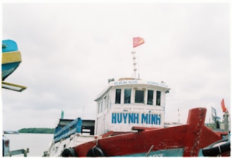 A boat with the name 'HUYNH MINH' painted in blue on its front is shown. On top of the boat is a red flag with a yellow star, indicating affiliation with Vietnam. The vessel is docked or stationary near a calm waterbody, with other boats and a bit of land visible in the background under a cloudy sky.