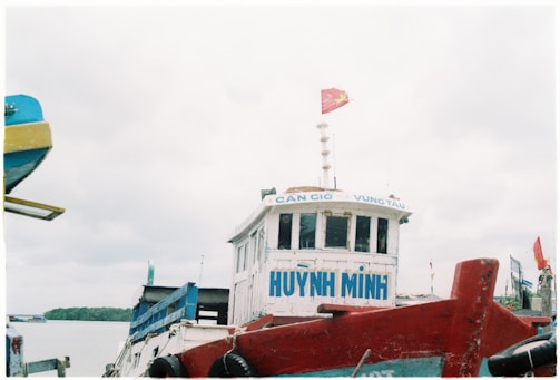 A boat with the name 'HUYNH MINH' painted in blue on its front is shown. On top of the boat is a red flag with a yellow star, indicating affiliation with Vietnam. The vessel is docked or stationary near a calm waterbody, with other boats and a bit of land visible in the background under a cloudy sky.