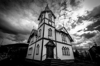 A black and white image of a gothic-style church with a tall steeple capped by a cross. The church features pointed arch windows and intricate woodwork, standing against a dramatic sky filled with turbulent, dark clouds.