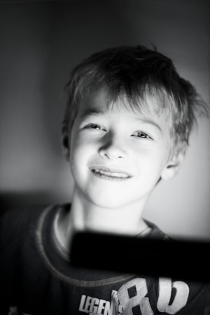 A young child with light-colored hair smiles gently, illuminated by soft lighting. The image is in black and white, focusing on the child's facial expression, which conveys a sense of innocence and happiness. The child's clothing is visible with some text partially visible across the chest.