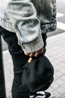 A person wearing a denim jacket and black jeans is seen holding a black cap by its strap. Part of the person's footwear, which appears to be sneakers, is visible. The background consists of a tiled floor and part of a street.