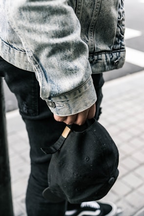 A person wearing a denim jacket and black jeans is seen holding a black cap by its strap. Part of the person's footwear, which appears to be sneakers, is visible. The background consists of a tiled floor and part of a street.
