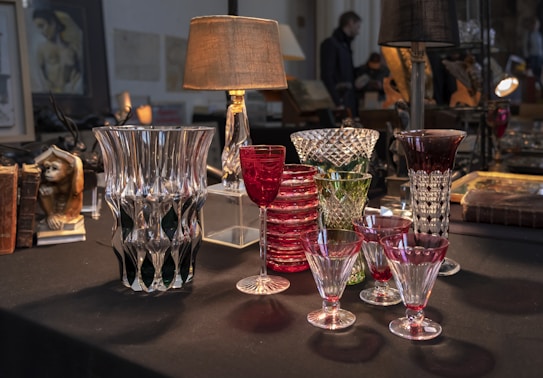 A collection of crystal glassware is displayed on a dark table. Various sizes and styles of ornate glasses include red goblets, green and clear glassware with intricate patterns. Behind the glassware, two lamps illuminate the scene, casting a warm glow. The background is filled with books, antique objects, and framed art, adding a vintage atmosphere.