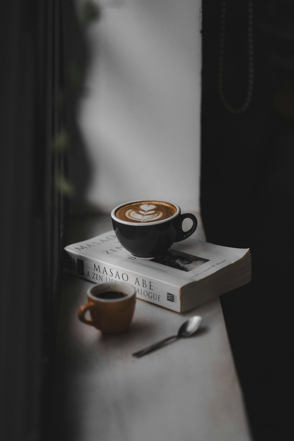 Latte gefüllte schwarze Teetasse auf der Oberseite des Buches in der Nähe der braunen Tasse am Fenster