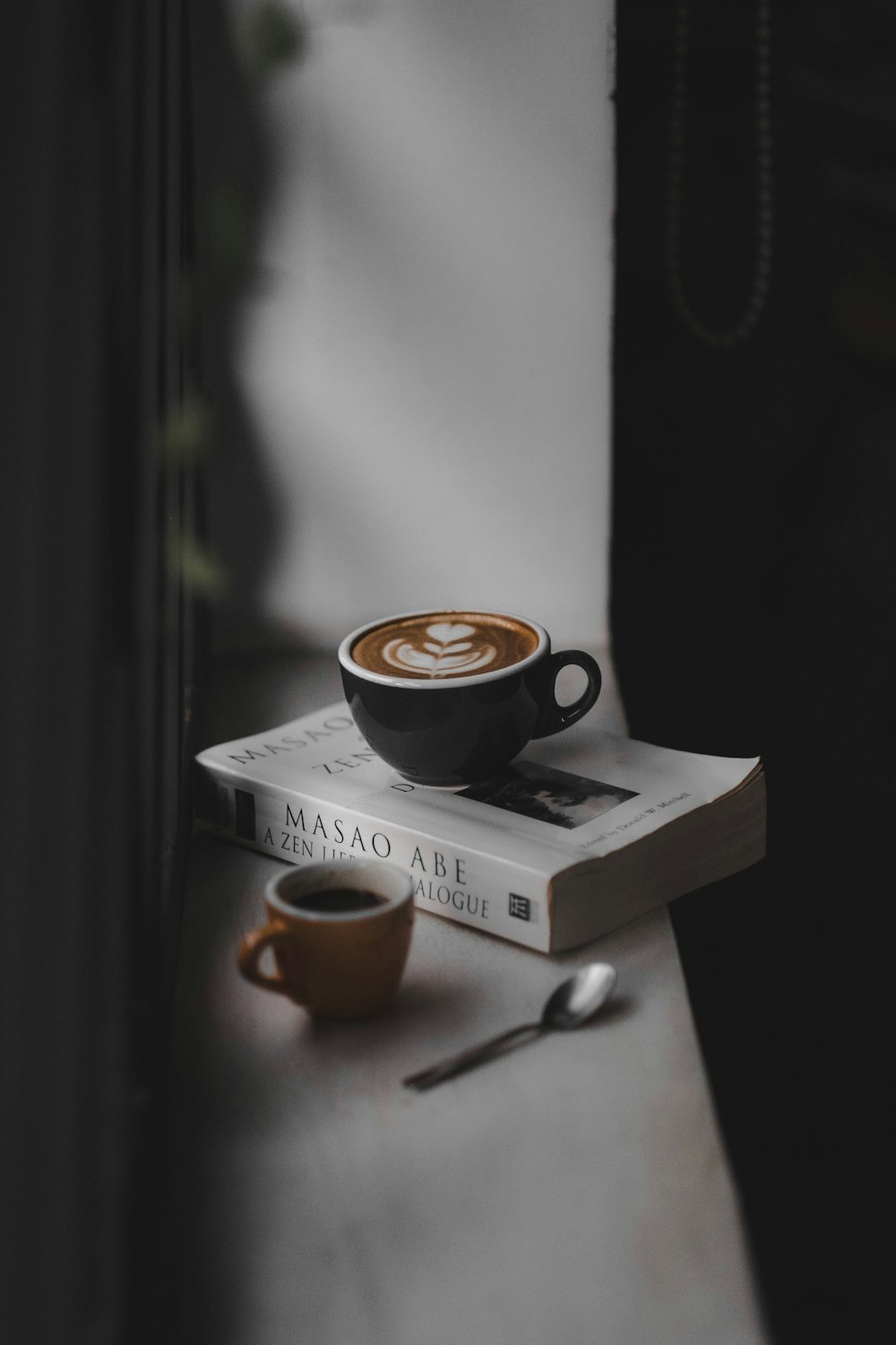 taza de té negro llena de café con leche en la parte superior del libro cerca de la taza marrón en la ventana