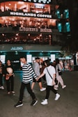 A group of people are walking on a city street in front of a multi-story building featuring signs for cafes and shops like Highlands Coffee and Legend Beer. The building is illuminated with warm light and people appear to be enjoying their evening.