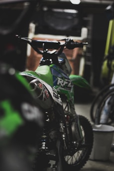 A green motocross bike is parked indoors, surrounded by dim lighting. The bike features detailed decals and has rugged tires suitable for off-road riding. Other bikes or parts can be partially seen in the blurred background, suggesting a garage or workshop setting.