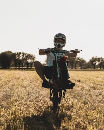 A person wearing a helmet performs a wheelie on a motorcycle in an open grassy field. The sunlight casts a warm hue over the scene, with trees visible in the background.