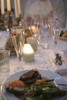 A beautifully set dinner table with a focus on a plate of food featuring meat, vegetables, and a slice of bread with an orange garnish. There are several glasses on the table, one containing a fizzy beverage with a raspberry. A lit candle and a napkin tied with a sprig of herbs add to the elegant setting.