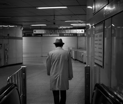 A person wearing a trench coat and a fedora hat is walking down an empty corridor in an underground metro station. The image is in black and white, giving it a classic, timeless feel. The background includes a shuttered gate and various signs in a foreign language, adding an element of mystery.