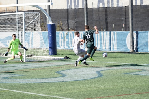 A soccer match is taking place on a green field. Two players are actively competing for the ball near the goal, with one player wearing a green jersey and the other in a white one. The goalkeeper, dressed in bright green, is positioned near the goal post, ready to defend. The goal net and a sports banner are visible in the background.