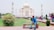 A man wearing a blue shirt and jeans is sitting on a bench in a garden area. The background features the stunning Taj Mahal, a famous white marble mausoleum with four minarets. There are people walking around and enjoying the tourist site.