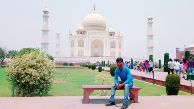 A man wearing a blue shirt and jeans is sitting on a bench in a garden area. The background features the stunning Taj Mahal, a famous white marble mausoleum with four minarets. There are people walking around and enjoying the tourist site.