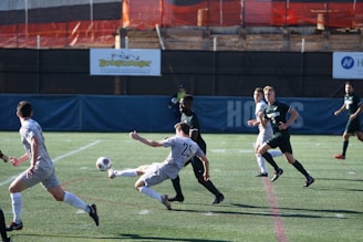 Two soccer teams are actively competing on a field. Players are in motion, with one player attempting to kick the ball. The background includes a fenced area with some industrial elements and advertising banners.