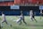 Two soccer teams are actively competing on a field. Players are in motion, with one player attempting to kick the ball. The background includes a fenced area with some industrial elements and advertising banners.