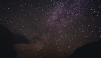 A vast night sky filled with countless stars, offering a glimpse of the Milky Way Galaxy. The dark silhouettes of mountains frame the bottom of the scene, creating a sense of depth and contrast. The stars vary in brightness, with some areas appearing denser with celestial bodies, hinting at distant nebulae and cosmic clouds.