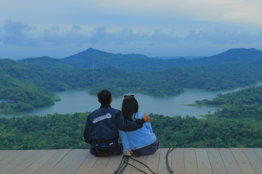 A couple sits together on a wooden platform overlooking a scenic view of a lush green landscape with rolling hills and a large tranquil lake. The sky is overcast with soft clouds, adding to the serene atmosphere.
