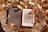 An e-reader with a low battery warning displayed on its screen is placed on a tree stump, covered with colorful autumn leaves. The e-reader has a cover, part of which is opened to reveal the screen.