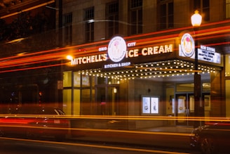 An ice cream shop with a vintage-style marquee sign illuminated at night. The sign features the name 'Mitchell's Ice Cream' with bright bulbs outlining the marquee. Light trails from passing vehicles create dynamic streaks of red and yellow across the image.