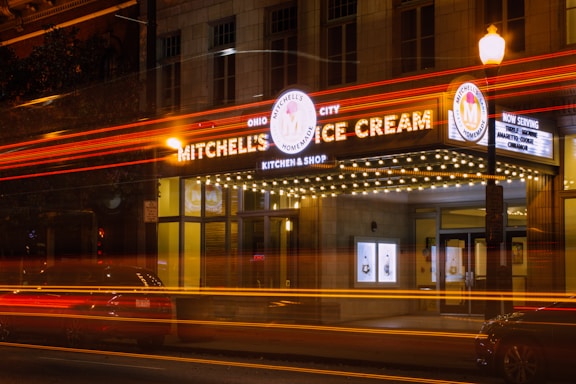 An ice cream shop with a vintage-style marquee sign illuminated at night. The sign features the name 'Mitchell's Ice Cream' with bright bulbs outlining the marquee. Light trails from passing vehicles create dynamic streaks of red and yellow across the image.