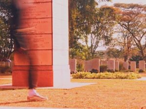 A person walks briskly past a red stone monument inscribed with names, located in a cemetery. The background reveals various gravestones amidst lush greenery and tall trees. The sunlight casts a warm glow over the scene, emphasizing the earthy tones of the grass.