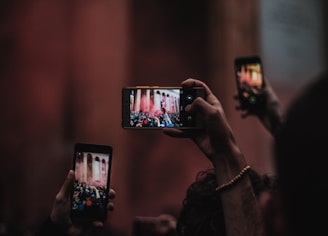 Several hands hold up smartphones capturing an event viewed through their screens. The focus is on the phone screens displaying a crowded scene in front of a large, columned structure with smoke or mist in the air. The background is blurred, emphasizing the action captured by the phones.