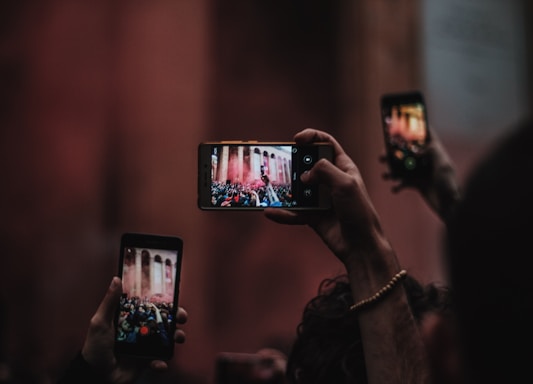 Several hands hold up smartphones capturing an event viewed through their screens. The focus is on the phone screens displaying a crowded scene in front of a large, columned structure with smoke or mist in the air. The background is blurred, emphasizing the action captured by the phones.