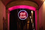 A neon sign hanging from an overhead archway in an alleyway, glowing with vibrant pink and blue colors. The sign has the words 'vinyl store' in the center, casting a warm light onto the surrounding walls and pavement, creating a cozy and inviting atmosphere.