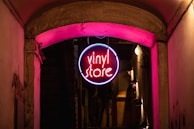 A neon sign hanging from an overhead archway in an alleyway, glowing with vibrant pink and blue colors. The sign has the words 'vinyl store' in the center, casting a warm light onto the surrounding walls and pavement, creating a cozy and inviting atmosphere.