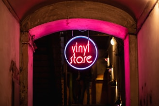 A neon sign hanging from an overhead archway in an alleyway, glowing with vibrant pink and blue colors. The sign has the words 'vinyl store' in the center, casting a warm light onto the surrounding walls and pavement, creating a cozy and inviting atmosphere.