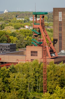 A large industrial structure with a distinctive red and green color scheme rises amidst a lush green landscape. The structure appears to be a mining or industrial tower, with visible ladders and platforms. Surrounding the structure are dense clusters of trees, and in the background, there is a clear sky with wind turbines visible on the horizon.