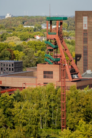 A large industrial structure with a distinctive red and green color scheme rises amidst a lush green landscape. The structure appears to be a mining or industrial tower, with visible ladders and platforms. Surrounding the structure are dense clusters of trees, and in the background, there is a clear sky with wind turbines visible on the horizon.