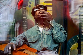 A person wearing a denim shirt and a brown cap is seated while holding a guitar with one hand. The individual is positioned in front of a colorful mural that includes keyboard motifs. The background features artistic and abstract elements with vibrant colors.