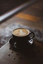 A cup of coffee with intricate latte art is placed on a wooden table, surrounded by scattered coffee beans. The lighting is soft, creating a cozy and inviting atmosphere.