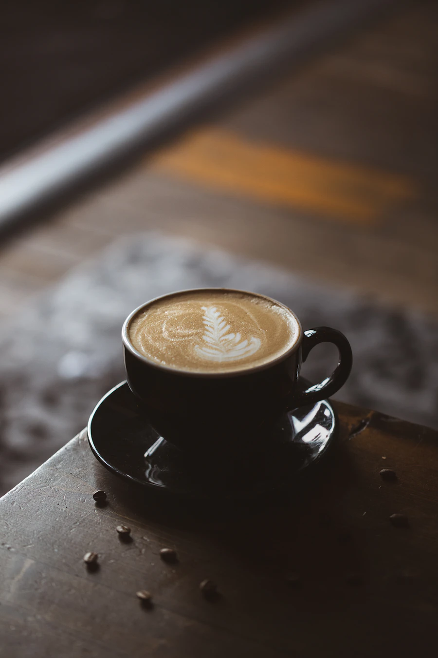 A dark cup of coffee with a poured rosetta on a scratched metal counter, loose beans scattered around it.