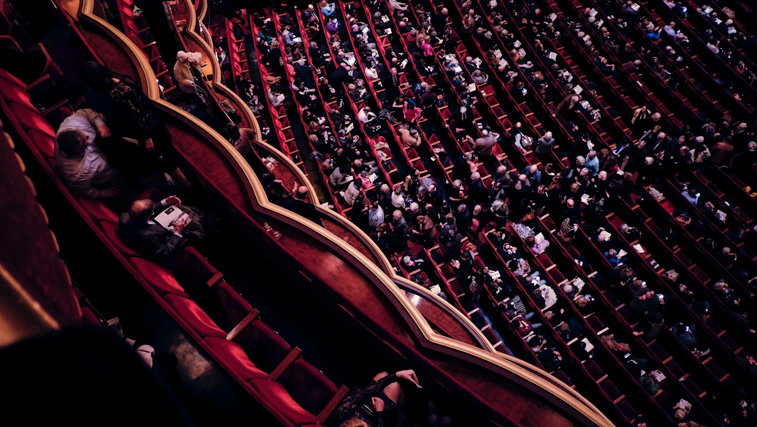 A theatre balcony photographed from above, rows of red seats curving away with the audience in them