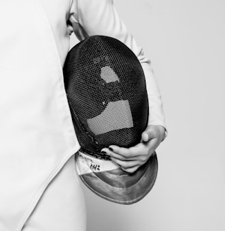 person wearing white fencing suit holding black mask inside room