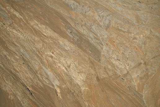 The image captures a close-up view of a rocky terrain with rugged, textured surfaces. The rocks seem to have a variety of shades, from light brown to dark gray, and display natural patterns and formations. Sparse patches of vegetation appear in some areas.