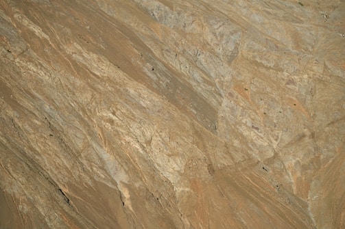 The image captures a close-up view of a rocky terrain with rugged, textured surfaces. The rocks seem to have a variety of shades, from light brown to dark gray, and display natural patterns and formations. Sparse patches of vegetation appear in some areas.