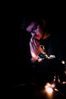 A person with glasses is seen in a dark setting, holding their hands in a praying position. Their face is softly illuminated by a string of fairy lights wrapped around their wrists, creating a tranquil and introspective atmosphere. The word 'JESUS' is visible on the sleeve of their clothing, adding a religious context to the scene.